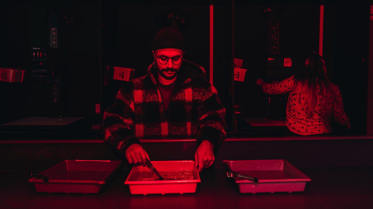Två personer står i mörkerrum med rödbelysning. Dem ena står vid en maskin och den andra doppar ett foto i en balja med vätska. Foto. 