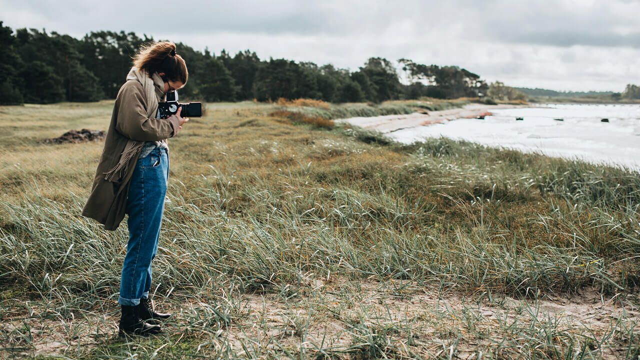 Utomhus, höst, på en gräsbevuxen sandstrand. En person står med sin kamera riktad ut mot havet. Foto. 
