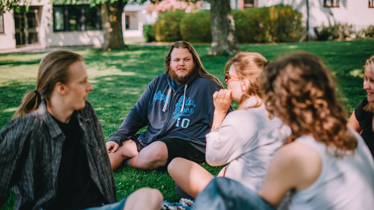 En grupp samtalande människor sitter på en gräsmatta. Foto. 